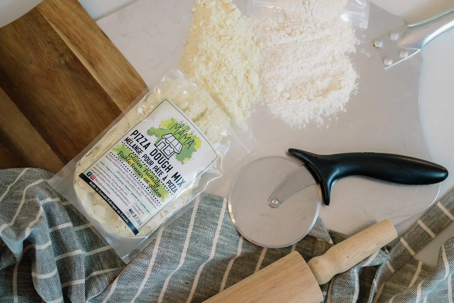Overview picture of the Dough Mama Gluten-Friendly Pizza Dough Mix package tipped over with mix spilling out onto a white kitchen counter with a bamboo cutting board, blue tea towel and black and silver pizza cutter