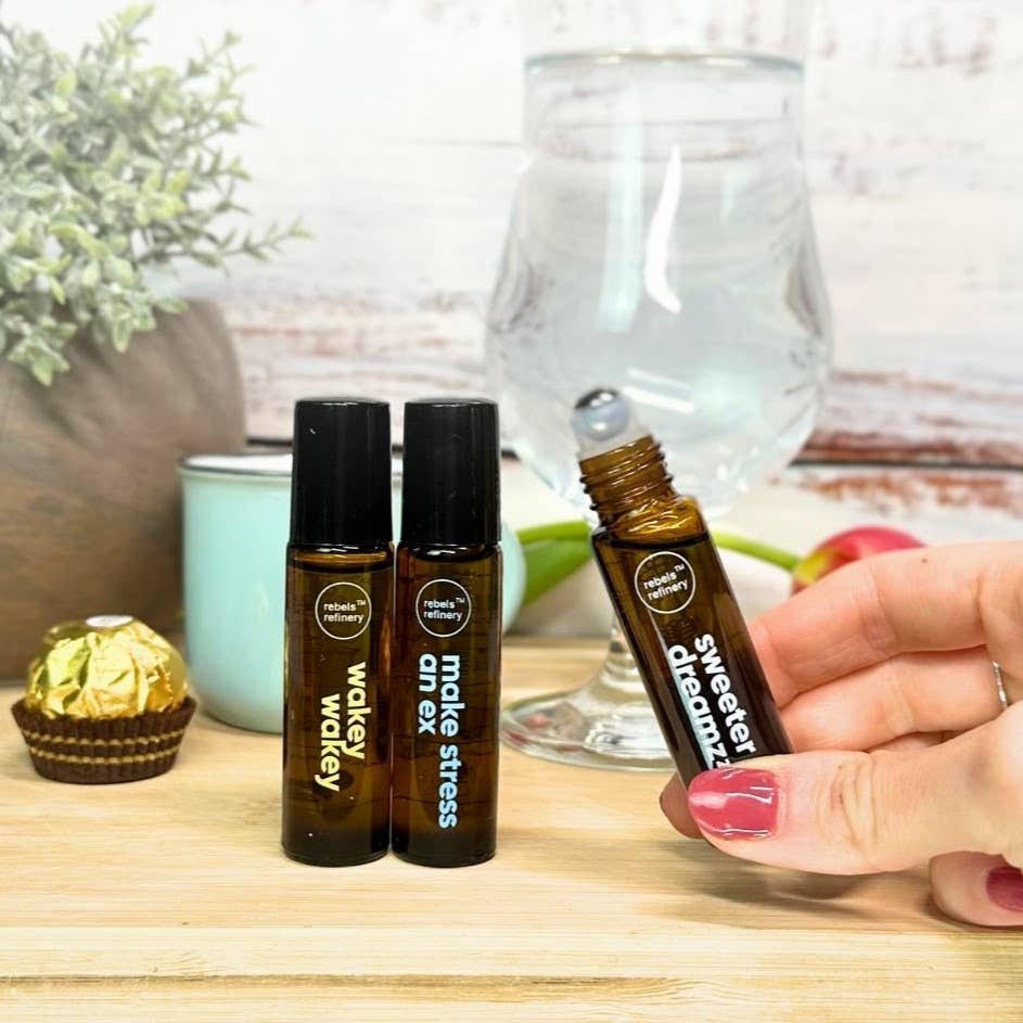 rebel refineries essential oil roller bottles with labels on a wooden surface, one being held by a hand.