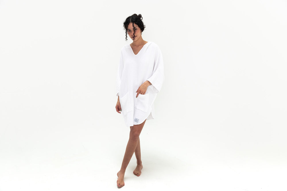 Tofino Towel cocoon surf poncho woman wearing white surf poncho hand in pocket she has darker skin dark hair up and bun white background very beautiful thin legs