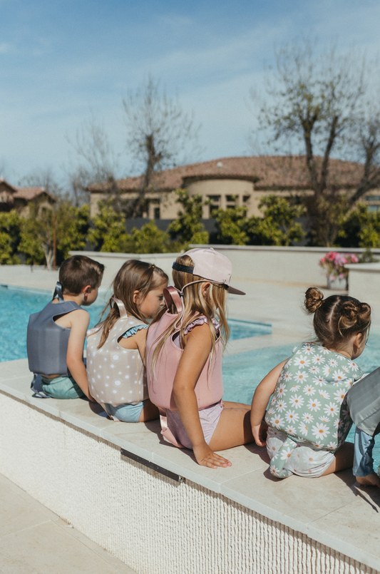 Swimming float vest picture showing 4 kids sitting poolside  all wearing their swim float vest one is wearing a current tyed SnapBack hat as well