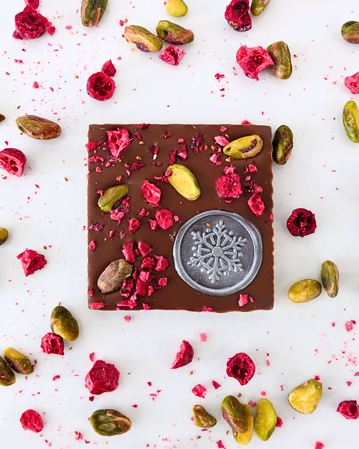 Elegant milk chocolate square topped with pistachios and crushed raspberries, featuring a silver snowflake imprint — festive gourmet chocolate available at The County Emporium, Carrying Place Ontario