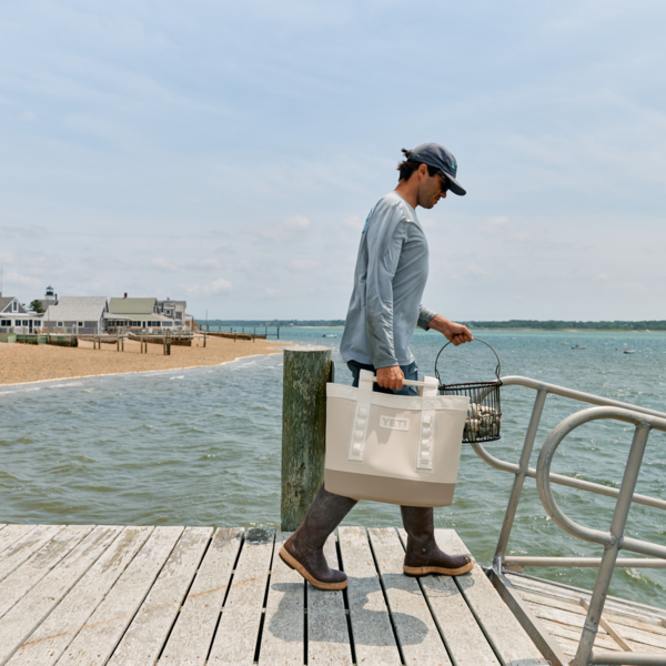 Man walking on a dock with a YETI beach tote and wire basket, coastal cottage views and ocean in the background – perfect for summer clamming, fishing, or beach adventures.