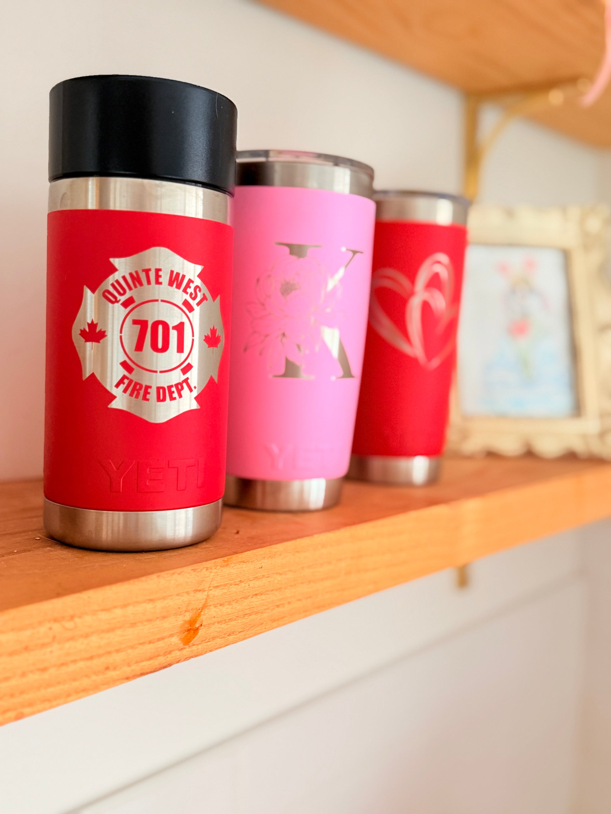 Custom engraved YETI tumblers in red and pink featuring a fire department logo, floral monogram, and heart design displayed on a wooden shelf