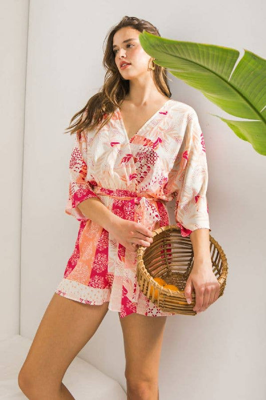 A printed woven romper featuring kimono top, elasticized waist with self sash tie, surplice back with tie and tassel detail